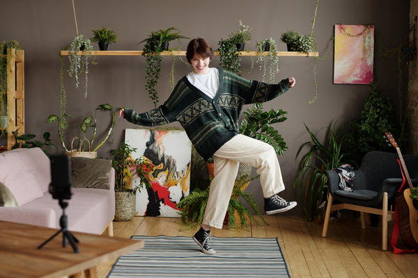 A girl with short dark hair dressed in a warm cardigan with patterns of light pants and sneakers smiles and dances filming herself on a phone that stands nearby on a tripod